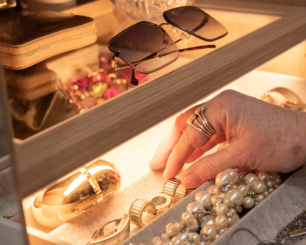 A hand with stacked silver rings reaching into a lit jewellery drawer of pearls and gold watches